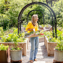 Woman standing in a garden with plants and a table, surrounded by greenery.